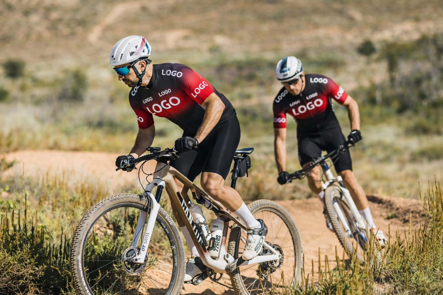 Two cyclists riding mountain bikes on a dirt trail with custom branding.